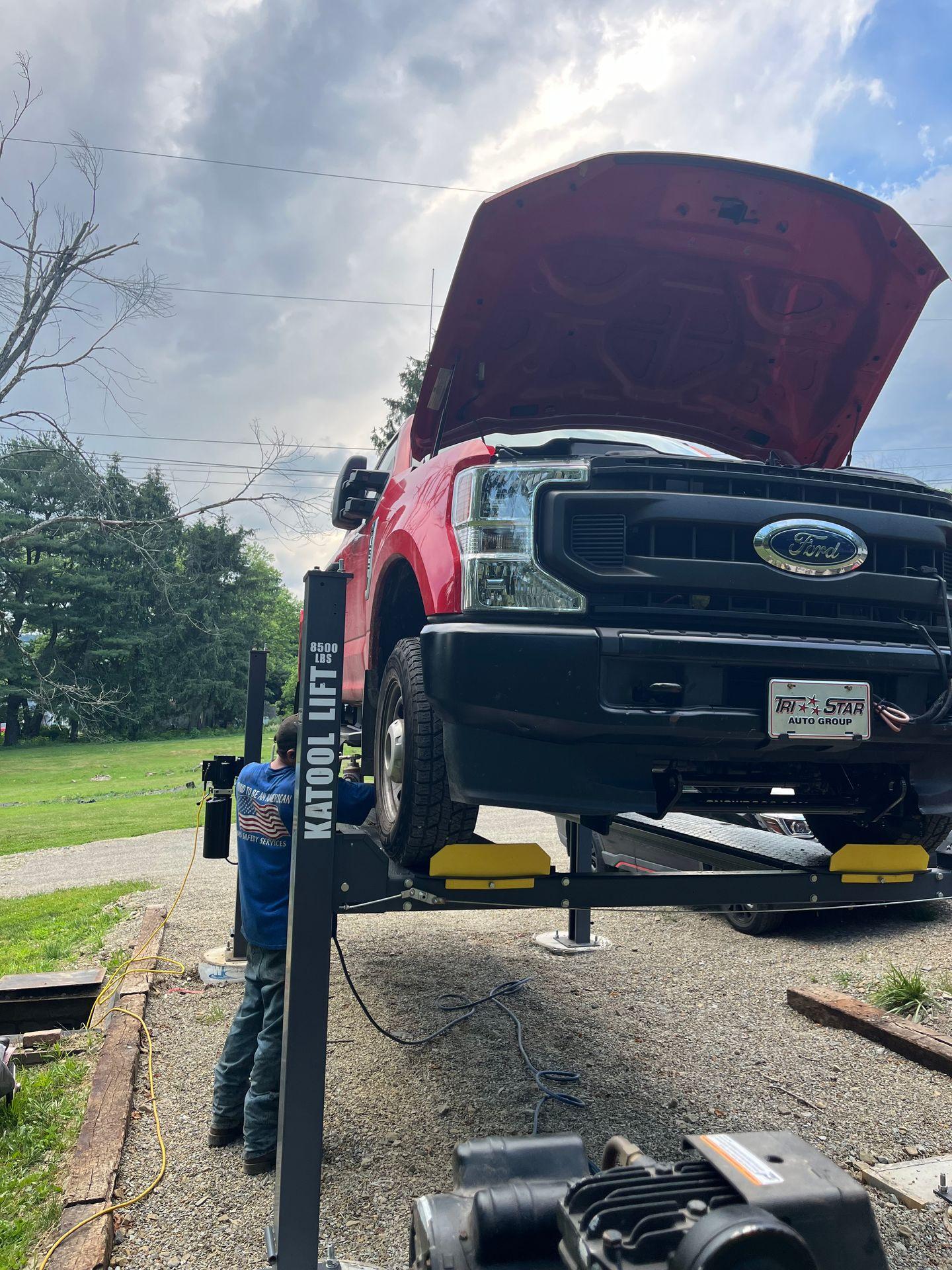 Mechanic working on a red pickup truck lifted on a hydraulic lift outdoors with the hood open.