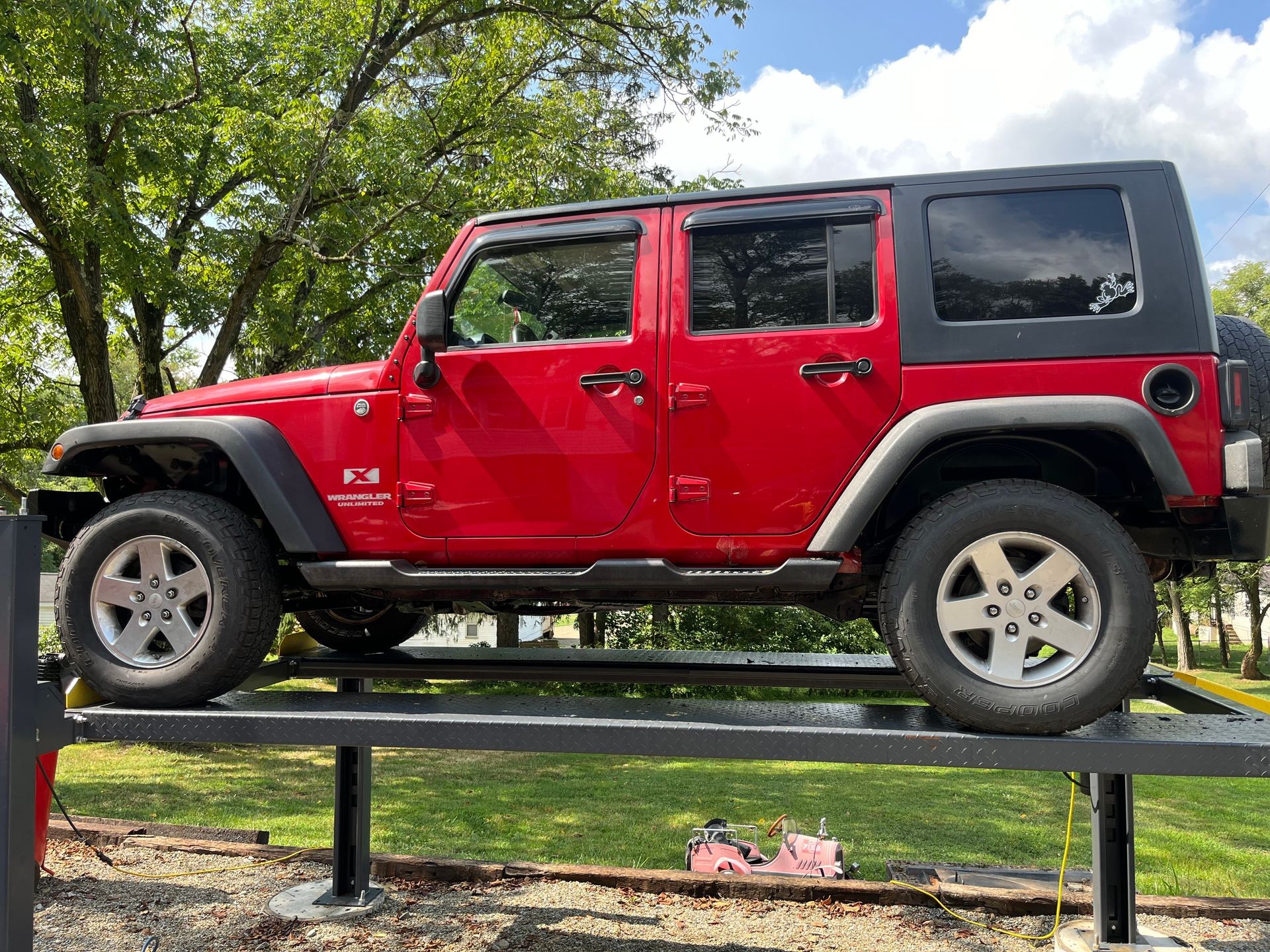 Red Jeep Wrangler Unlimited parked on an elevated platform with trees and cloudy sky in the background.