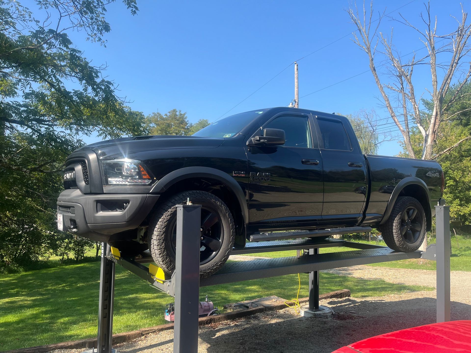 Black pickup truck elevated on a hydraulic lift outdoors, surrounded by trees and a clear blue sky.